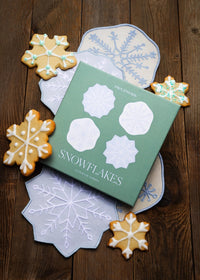 A green box labeled 'SNOWFLAKES' with white snowflake cutout designs, arranged on a dark wooden surface with decorated snowflake-shaped cookies and matching snowflake mats.