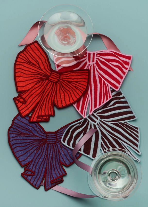 Four fabric bow cocktail napkins in red, pink-and-white striped, purple-and-dark-striped, and dark burgundy-and-white-striped patterns, arranged on a light blue background with two clear plastic discs.