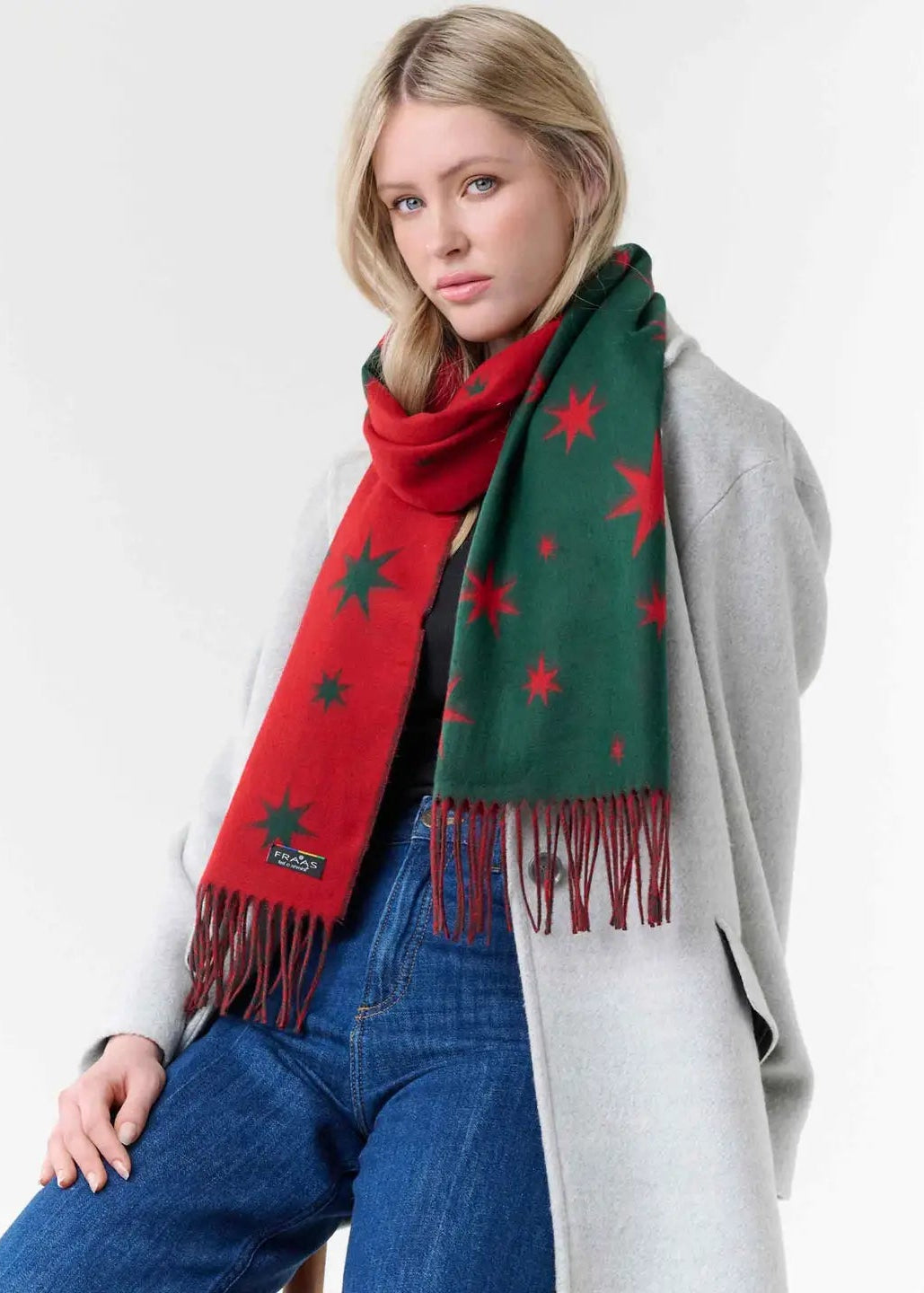 A woman wearing a festive Christmas-themed scarf with red and green sections and red star patterns, styled with a light gray coat and blue jeans against a plain white background.