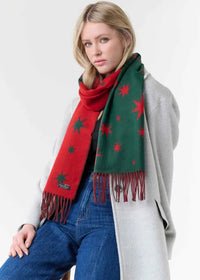 A woman wearing a festive Christmas-themed scarf with red and green sections and red star patterns, styled with a light gray coat and blue jeans against a plain white background.