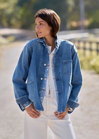 A woman wearing a light blue denim jacket over a white plaid shirt and white pants, standing outdoors on a sunlit path with a blurred natural background.