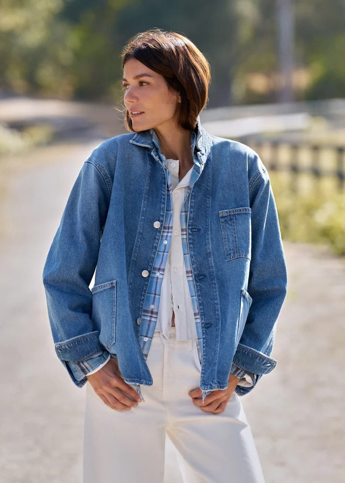 A woman wearing a light blue denim jacket over a white plaid shirt and white pants, standing outdoors on a sunlit path with a blurred natural background.