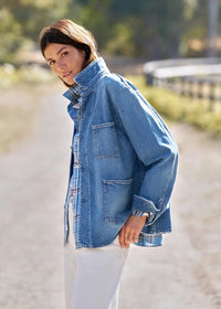 A woman wearing a light blue denim jacket over a plaid shirt and white pants, standing on a sunlit dirt path outdoors.
