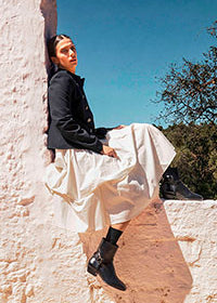 A model wearing a black jacket, a voluminous white skirt, and black boots sits on a textured stucco wall against a clear blue sky.