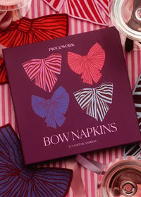 A square box of Pieceworks cocktail napkins lies on a pink-and-white striped fabric, surrounded by folded napkins and two wine glasses. The box is deep purple with four bow illustrations in pink, red, blue, and white striped patterns, and the text 'BOW NAPKINS' is printed on the front.