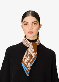 A woman with dark hair pulled back wears a black knit sweater and a patterned silk scarf. The scarf features a brown and beige botanical design with bright blue edges, shown against a plain light gray background.