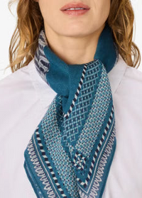 A close-up of a model wearing a teal and navy blue patterned scarf tied around their neck, paired with a white collared shirt against a clean white background.