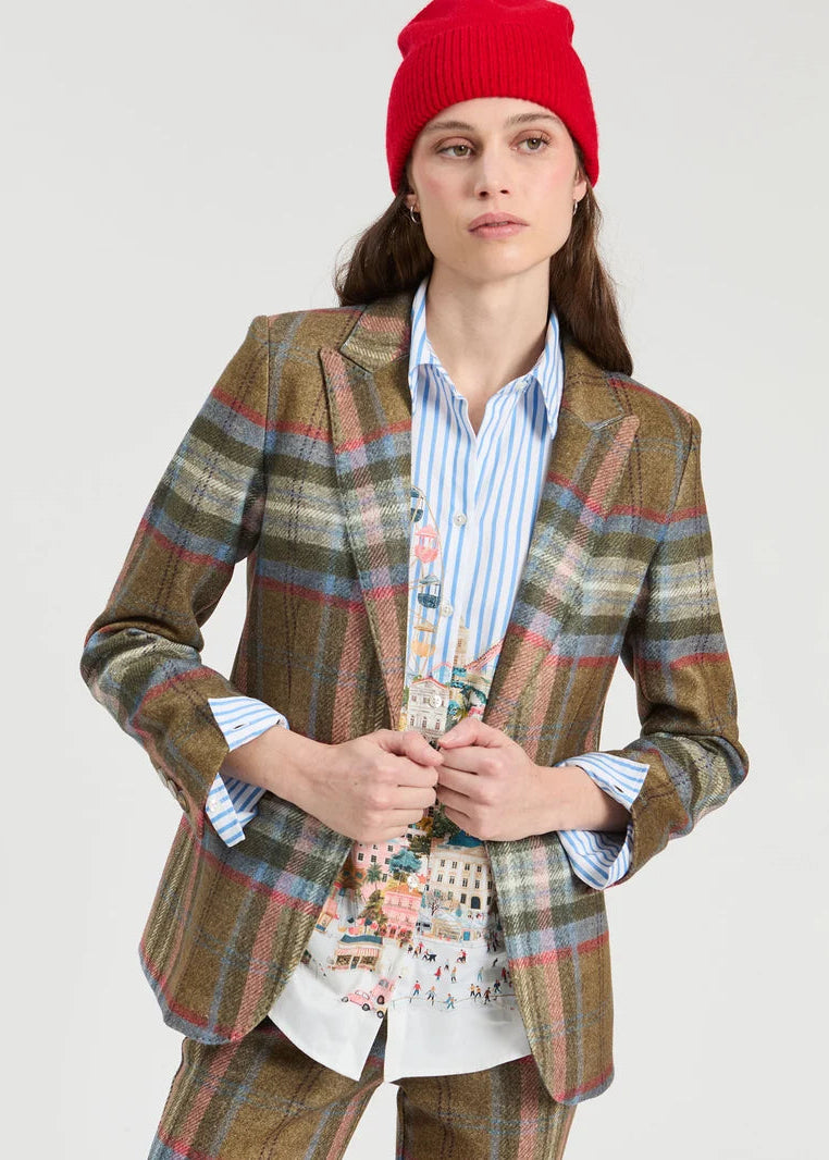 A model wearing a brown plaid blazer and matching trousers over a blue and white striped shirt with a colorful graphic print, accessorized with a bright red knit beanie, against a light gray studio background.