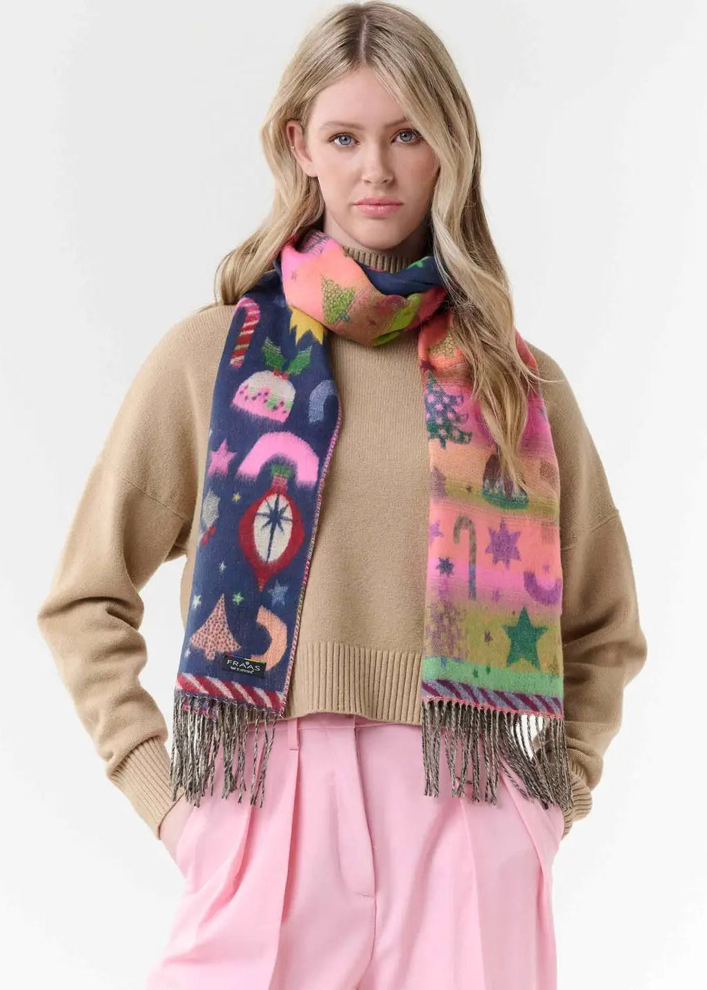 A woman modeling a vibrant, multicolored patterned scarf with fringe, styled over a beige sweater and pink trousers against a plain white background.