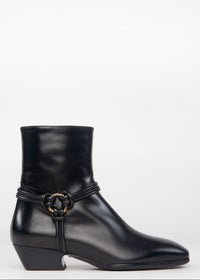 A black leather ankle boot with a low block heel and a decorative strap detail featuring a gold-tone ring buckle, shown against a plain white background.