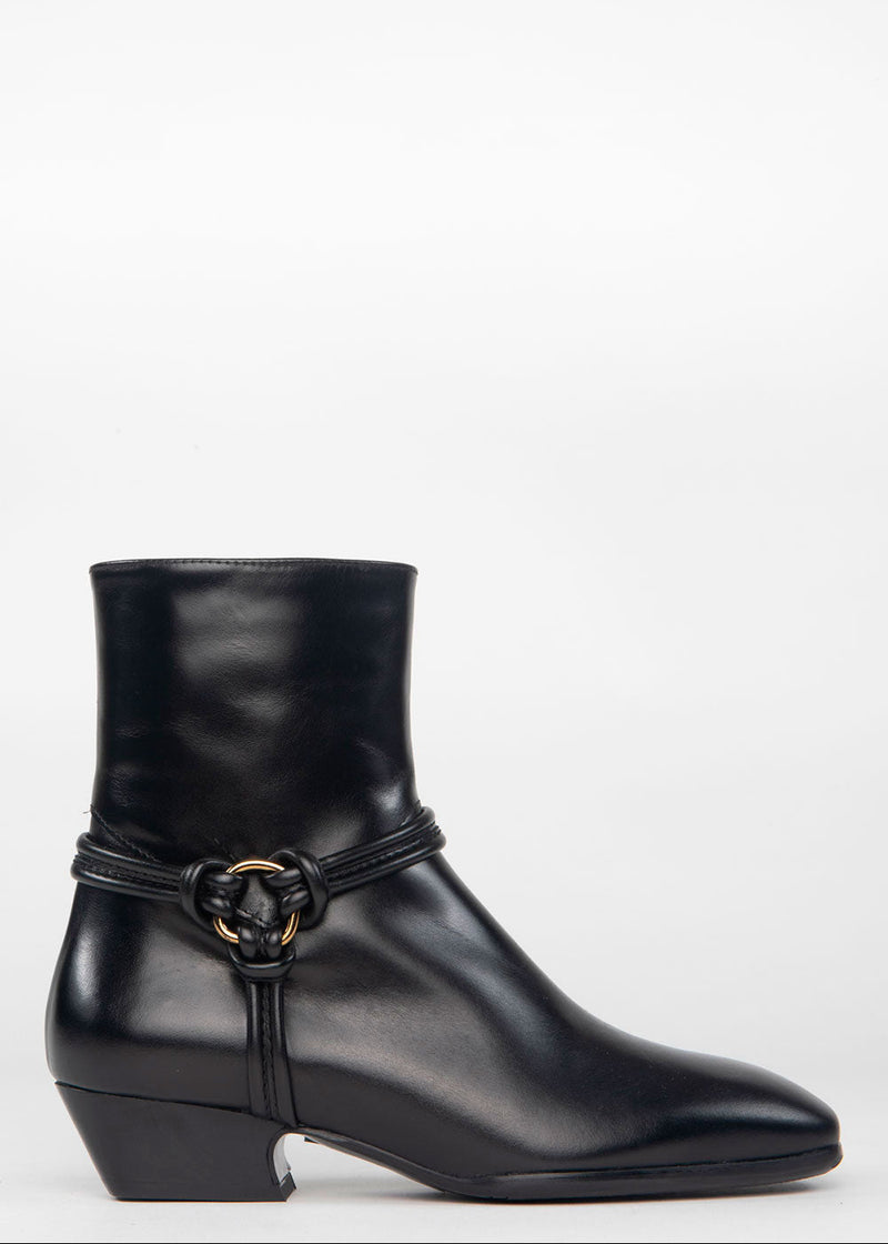 A black leather ankle boot with a low block heel and a decorative strap detail featuring a gold-tone ring buckle, shown against a plain white background.
