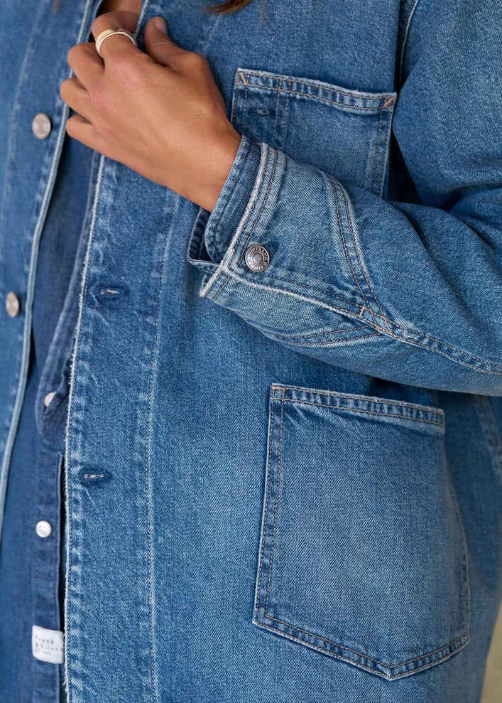 Close-up of a person wearing a medium-wash blue denim button-up shirt jacket with front pockets and a visible hand adjusting the collar.