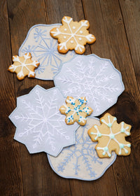 Four snowflake-shaped sugar cookies with white, blue, and green icing are arranged on top of four matching embroidered fabric coasters with white, light blue, and pale blue snowflake patterns, all set on a dark brown wooden table.