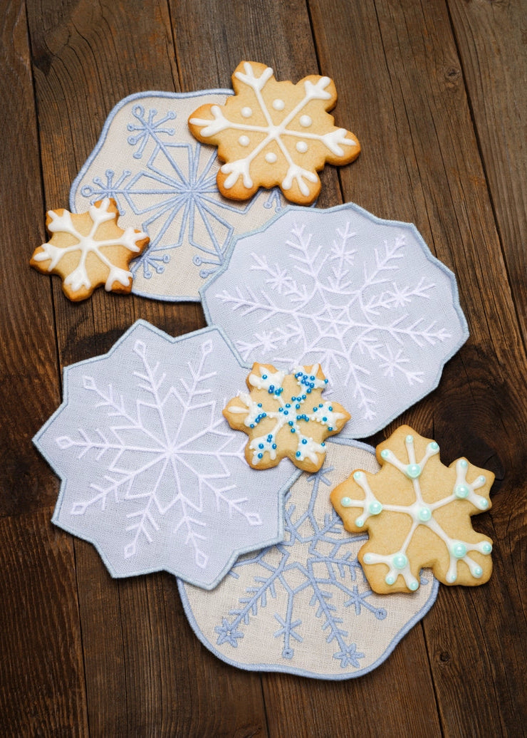 Four snowflake-shaped sugar cookies with white, blue, and green icing are arranged on top of four matching embroidered fabric coasters with white, light blue, and pale blue snowflake patterns, all set on a dark brown wooden table.