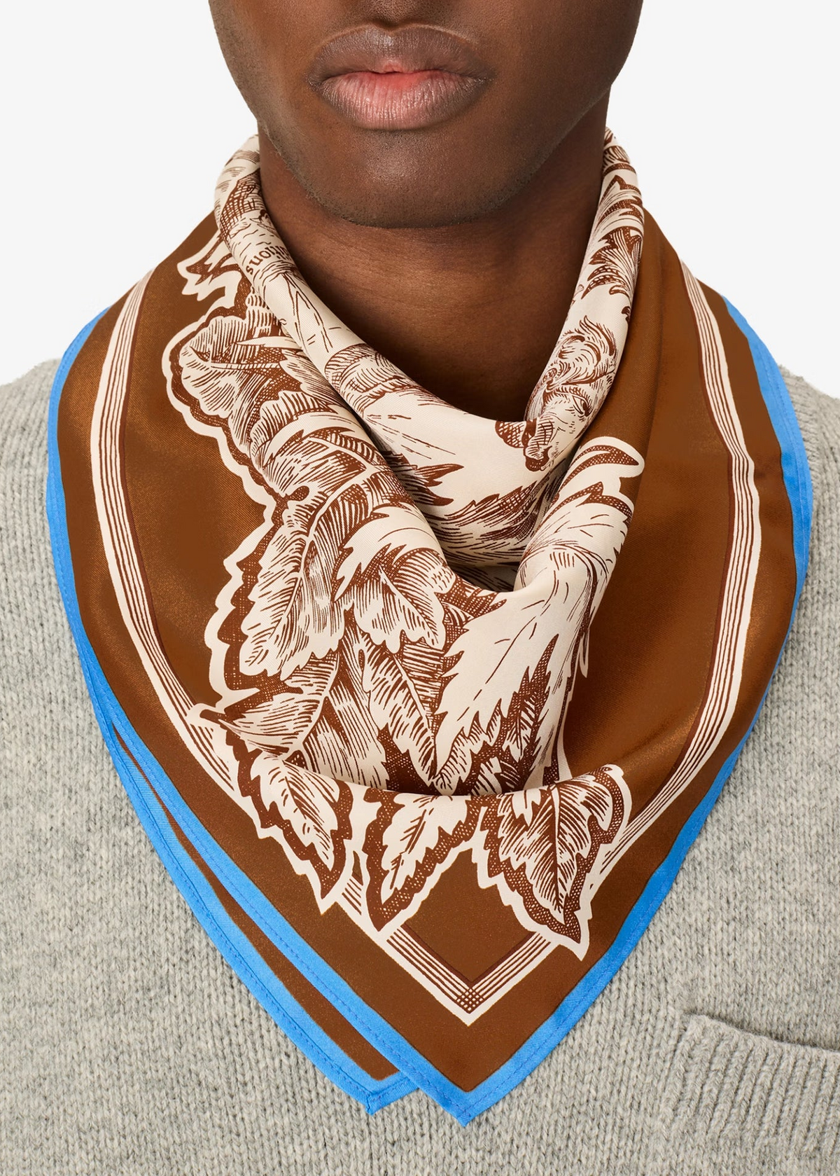 A close-up of a model wearing a brown and beige patterned silk scarf with a blue border, styled around the neck over a gray knit sweater.