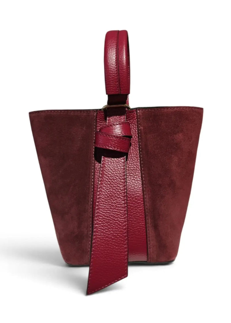 A structured leather bucket bag in deep burgundy, featuring a mix of smooth and pebbled textures, a top handle, and a hanging leather tie detail, shown on a clean white background.