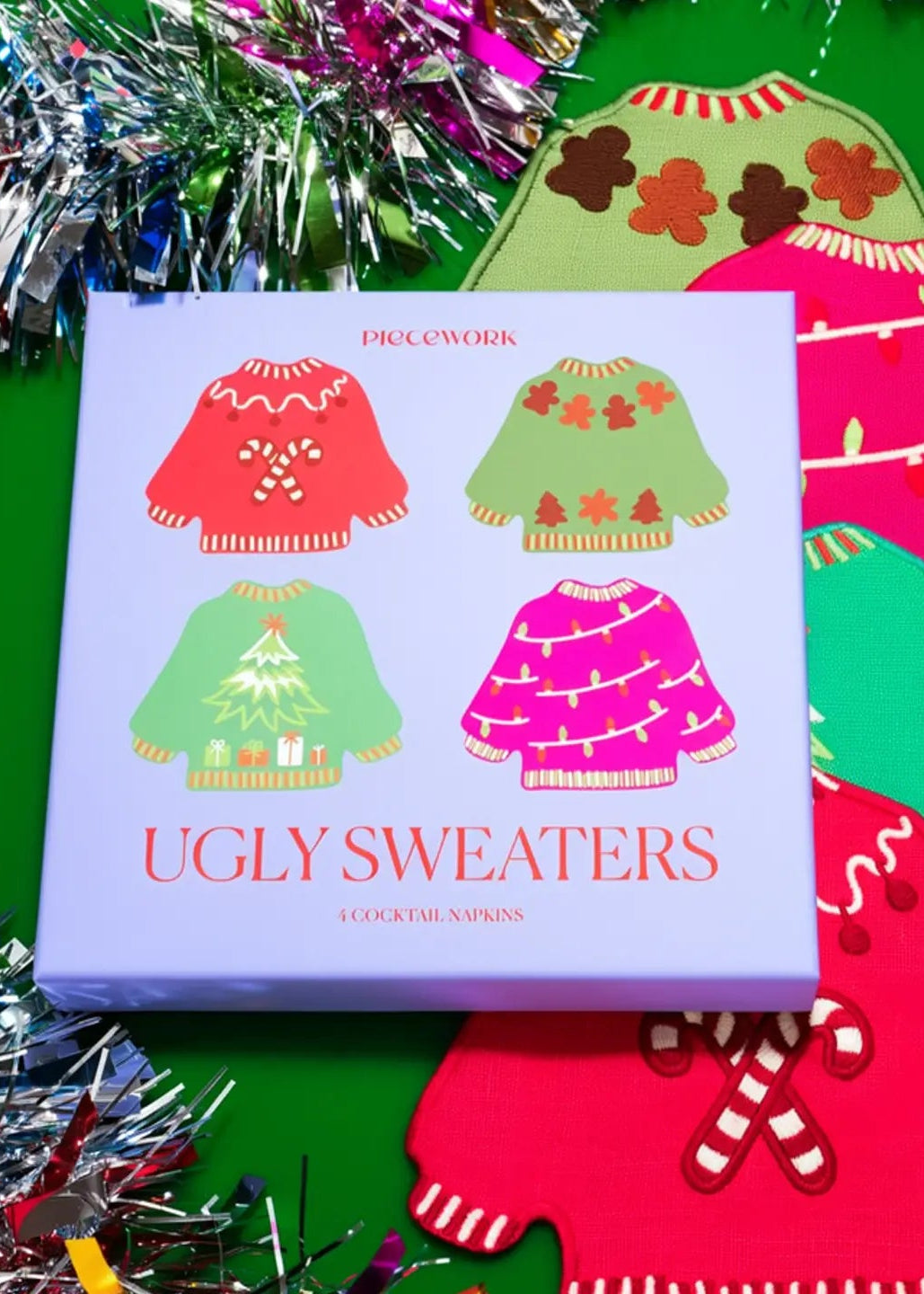 A square product box labeled 'Pieceworks' and 'UGLY SWEATERS' sits on a bright green surface with festive tinsel. The box shows four colorful sweater illustrations, and several matching sweater-shaped items are arranged around it in red, green, pink, and brown tones.