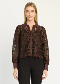 A woman modeling a brown sheer lace long-sleeve blouse with a dark underlayer, paired with black pants, against a plain light gray studio background.