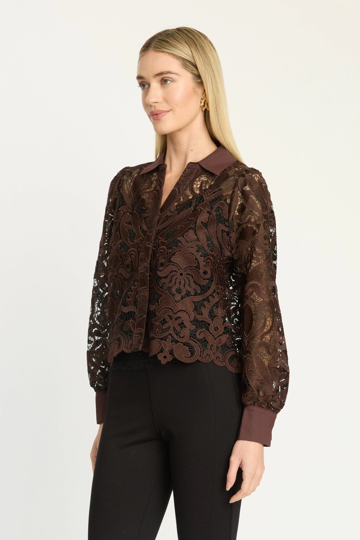 A woman modeling a brown lace long-sleeve blouse with a scalloped hem and dark brown collar, paired with black pants, against a plain light gray studio background.