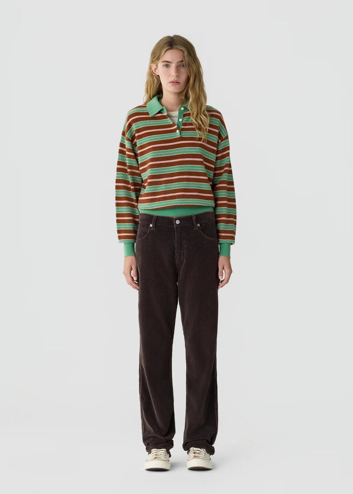 A model stands against a light gray studio background wearing a long-sleeve striped knit polo shirt with green, orange, and brown stripes, paired with dark brown corduroy pants and white sneakers.