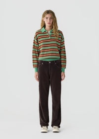 A model stands against a light gray studio background wearing a long-sleeve striped knit polo shirt with green, orange, and brown stripes, paired with dark brown corduroy pants and white sneakers.