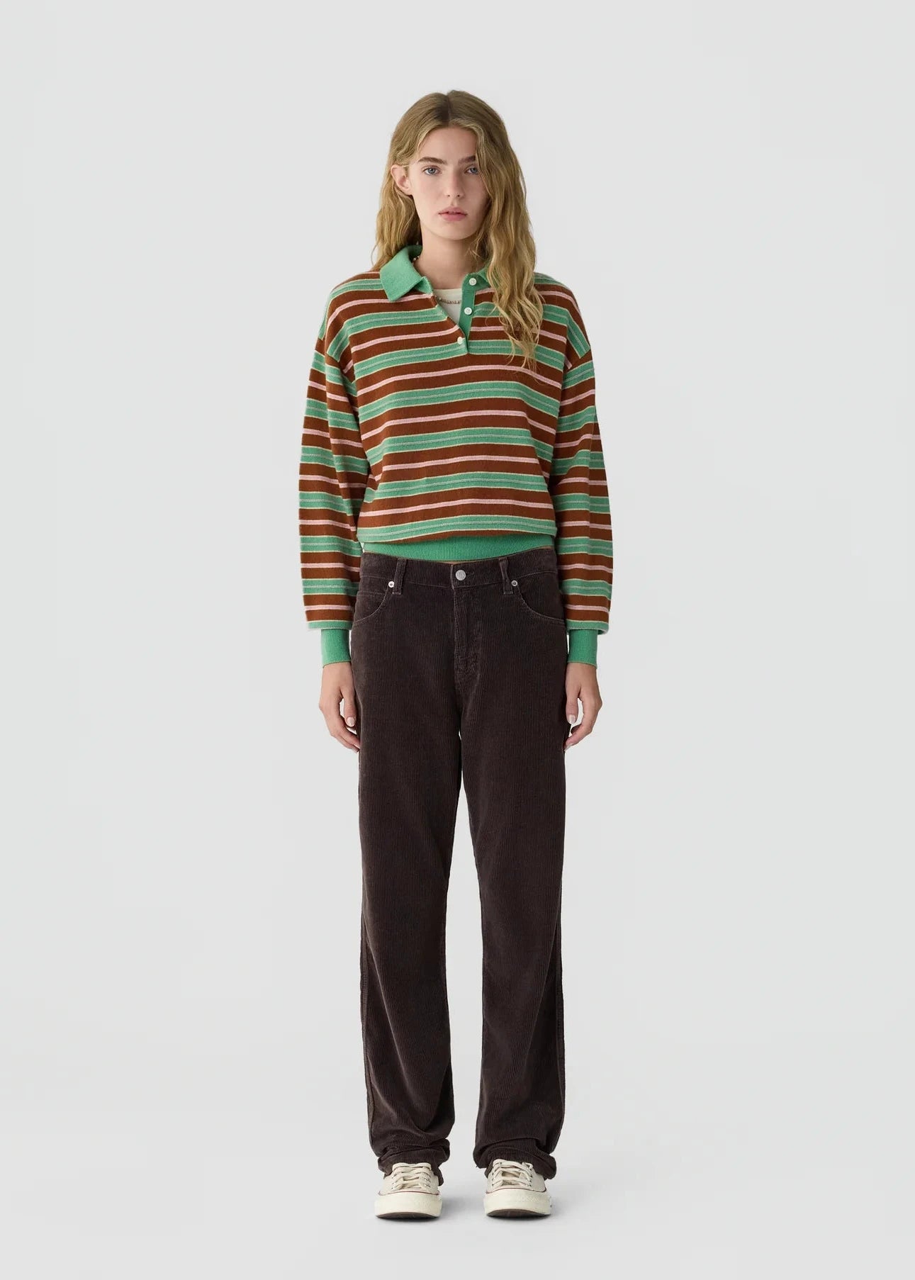 A model stands against a light gray studio background wearing a long-sleeve striped knit polo shirt with green, orange, and brown stripes, paired with dark brown corduroy pants and white sneakers.
