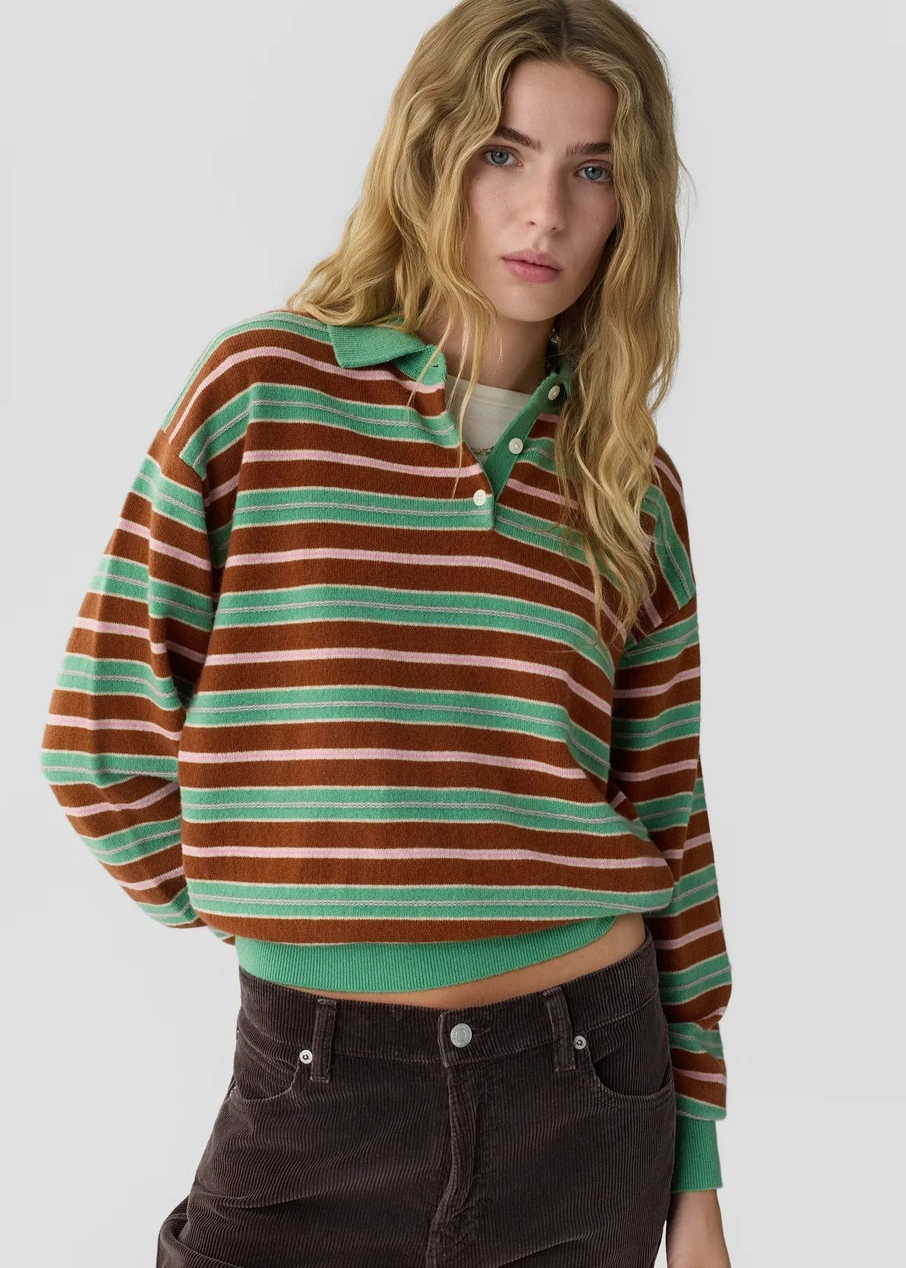 A woman wearing a long-sleeve knit polo sweater with horizontal brown, mint green, and cream stripes, paired with dark brown corduroy pants, against a light gray studio background.