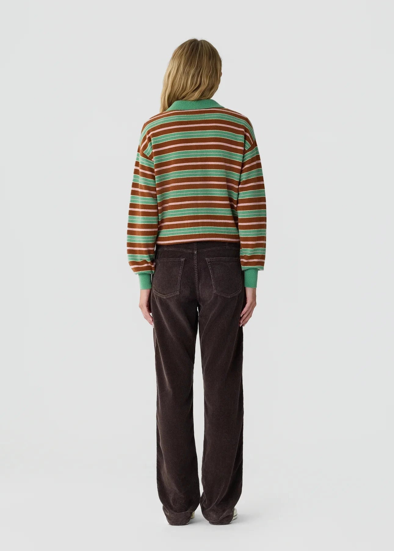 A model viewed from the back wearing a green, brown, and red striped knit sweater and dark brown corduroy trousers, set against a plain light gray studio background.