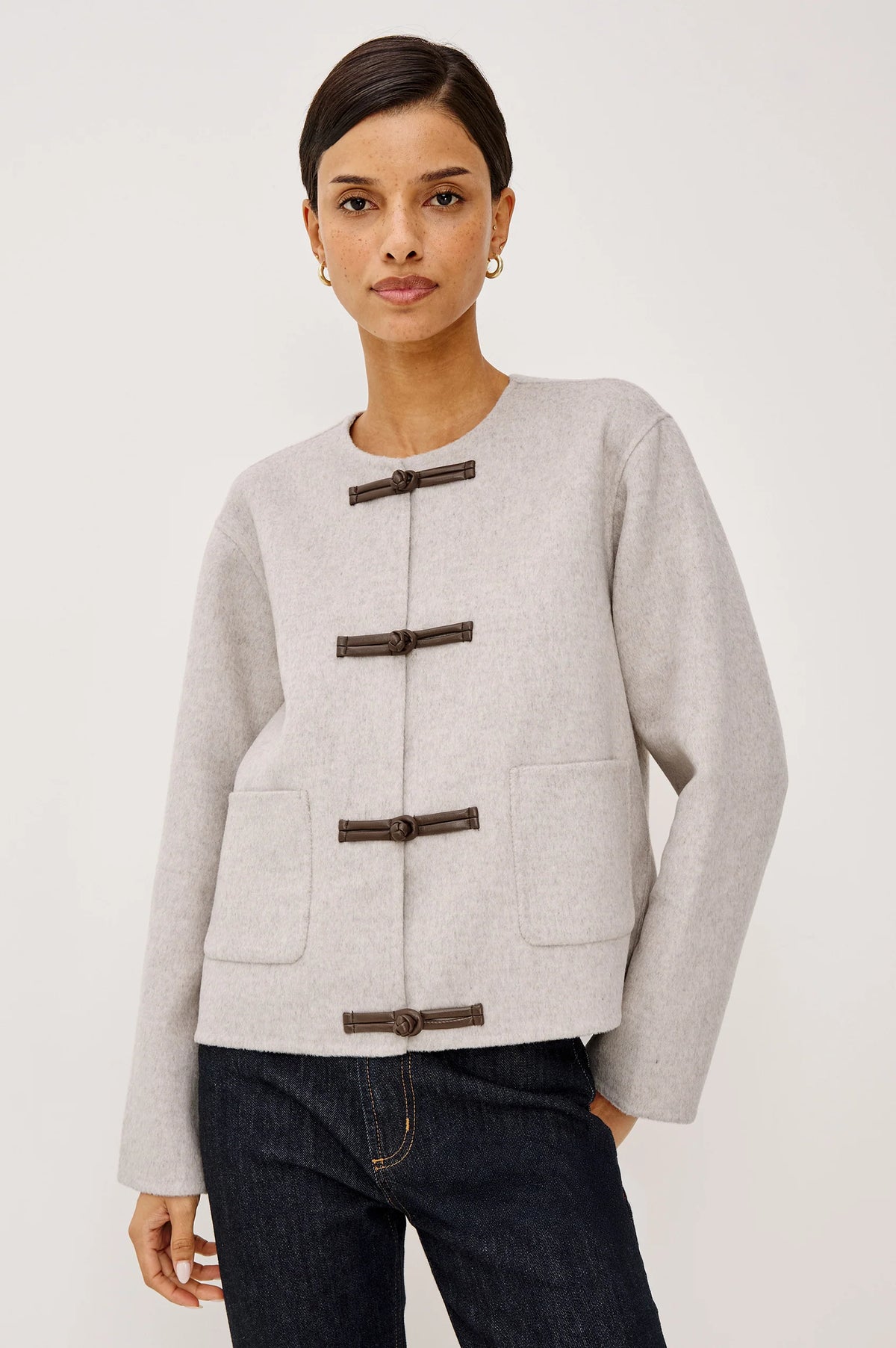 A woman modeling a light gray wool-blend jacket with dark brown toggle closures and front patch pockets, paired with dark blue jeans, against a plain light gray studio background.