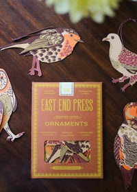 Box of East End Press ornaments with decorative paper birds on a wooden surface