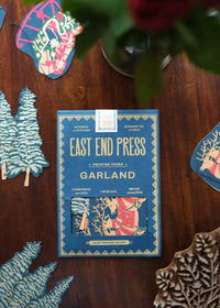 A boxed holiday garland by East End Press is displayed on a dark wooden surface, surrounded by matching decorative paper cutouts and a blurred plant in the background.