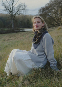 A woman sits in a grassy field outdoors, wearing a light gray knit sweater, a long white skirt, and a dark patterned scarf.