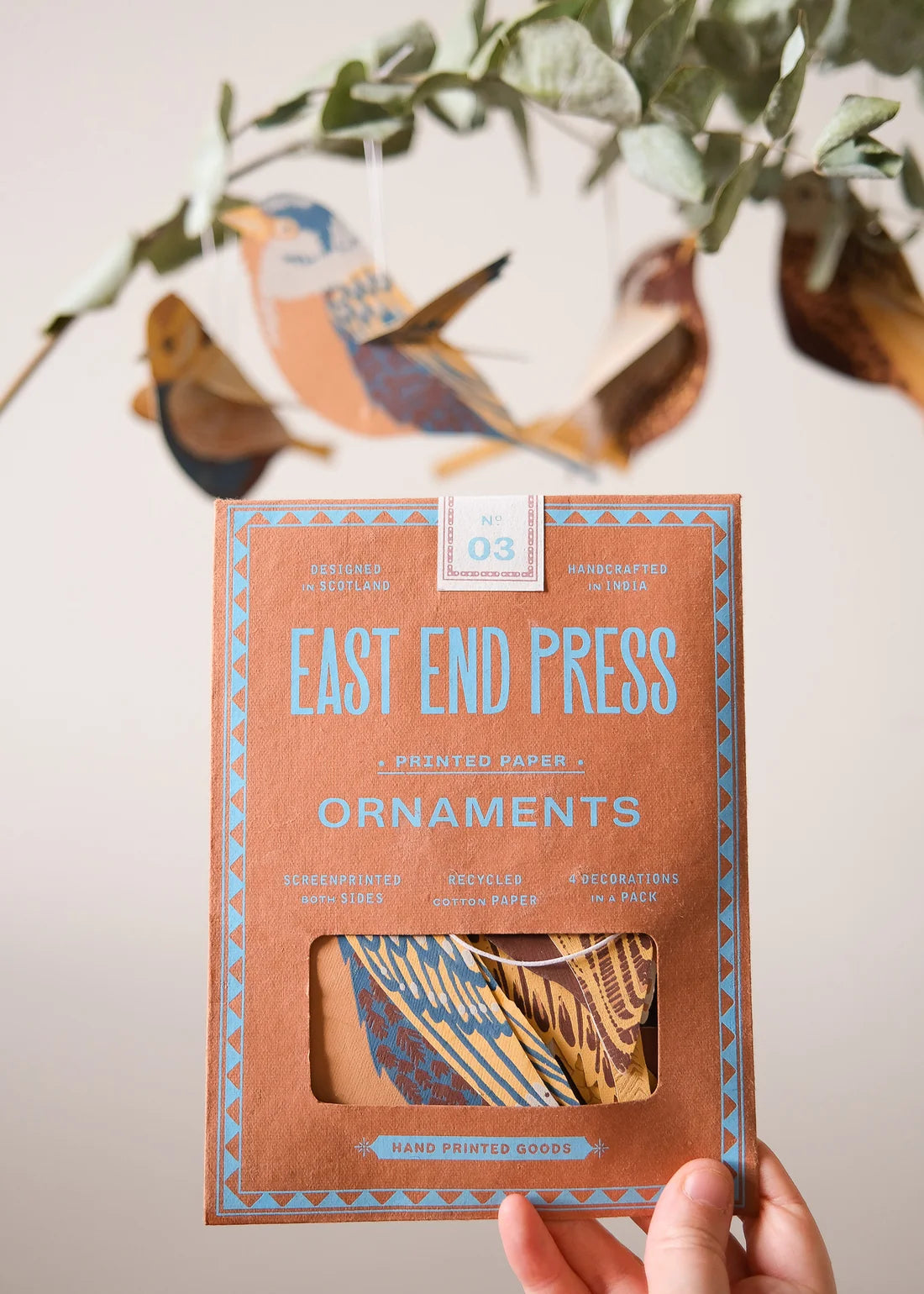 A hand holds a box of East End Press paper ornaments, showing the packaging with a window revealing patterned paper inside. The background is softly blurred with hanging bird ornaments and greenery.