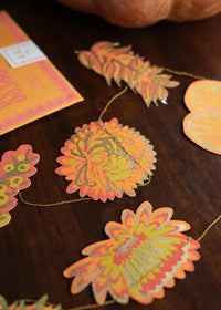 Autumn-themed paper garland with scalloped turmeric and maple leaf cutouts in orange, yellow, and green, displayed on a dark wooden tabletop next to a pumpkin and a matching orange card.