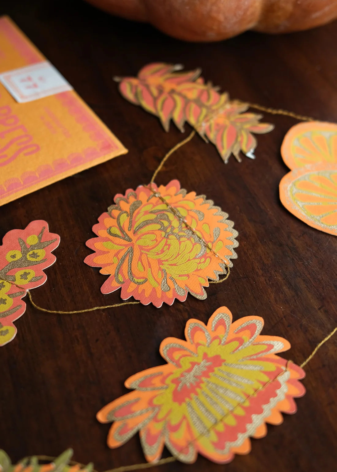 Autumn-themed paper garland with scalloped turmeric and maple leaf cutouts in orange, yellow, and green, displayed on a dark wooden tabletop next to a pumpkin and a matching orange card.