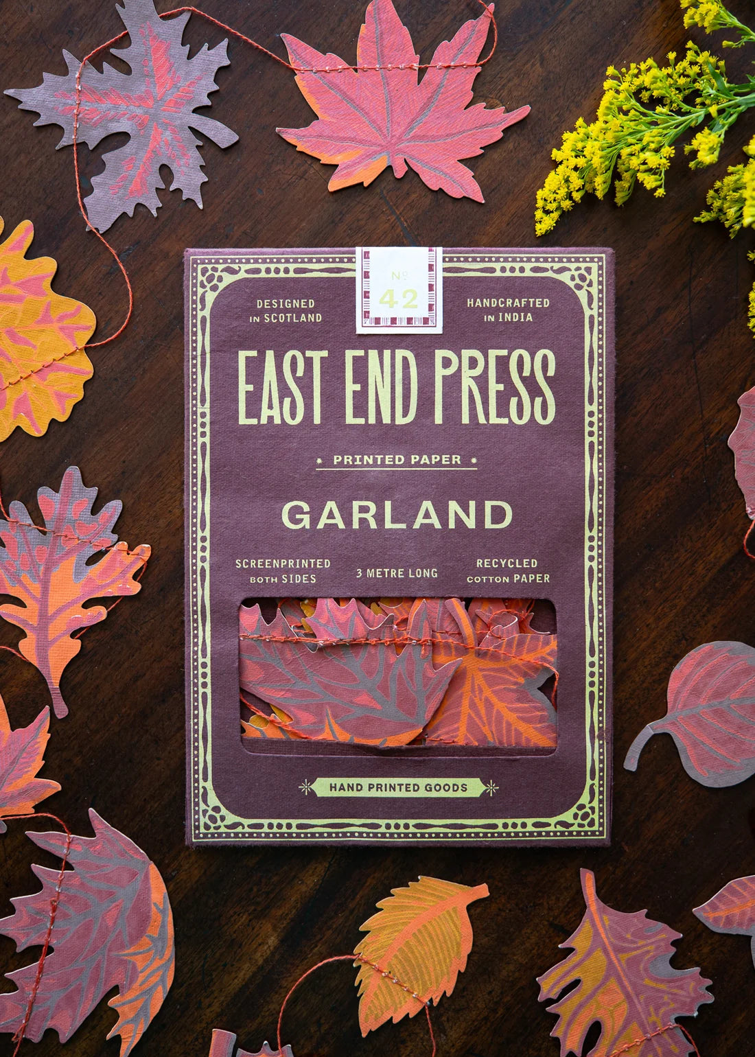 A package of East End Press Garland printed paper sheets lies on a dark wooden surface surrounded by colorful autumn leaves and small yellow flowers. The package is mauve-purple with cream text and border details, and a window near the bottom shows folded paper samples in pink, orange, and red tones.