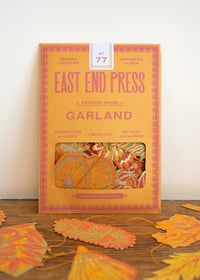 East End Press Garland No. 77 printed paper cookie cutters in a warm orange box, displayed on a wooden surface with several orange and yellow paper leaves scattered around.