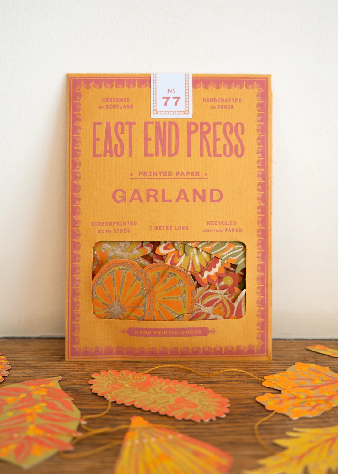East End Press Garland No. 77 printed paper cookie cutters in a warm orange box, displayed on a wooden surface with several orange and yellow paper leaves scattered around.