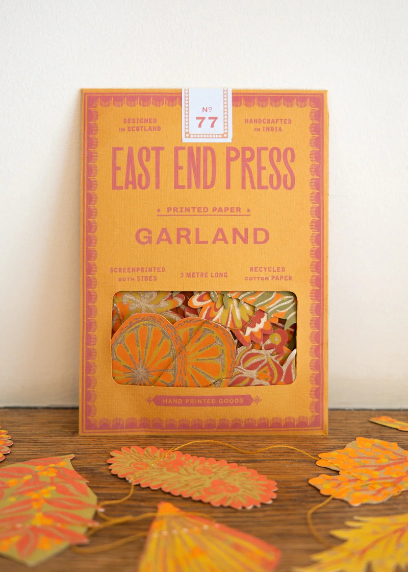 East End Press Garland No. 77 printed paper cookie cutters in a warm orange box, displayed on a wooden surface with several orange and yellow paper leaves scattered around.