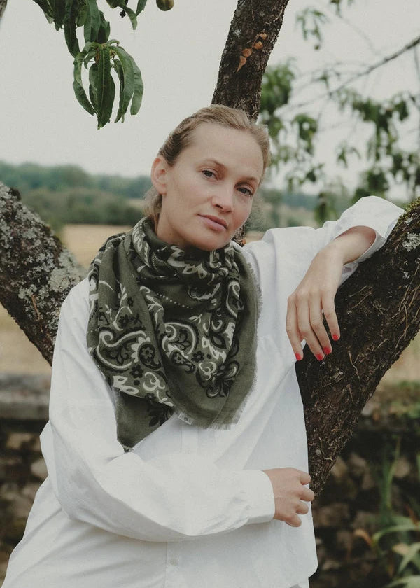 A woman wearing a dark olive green paisley bandana scarf and a white shirt, leaning against a tree trunk outdoors.