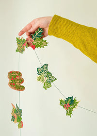 A hand wearing a yellow knit sweater holds a hanging Christmas garland made of green holly leaves and pinecones with small red berries against a plain light background.