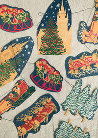 A festive Christmas garland made of multiple  paper cutouts on a string, featuring Santa in sleighs, Christmas trees, a castle, and holiday scenes, arranged on a light beige textured fabric background.