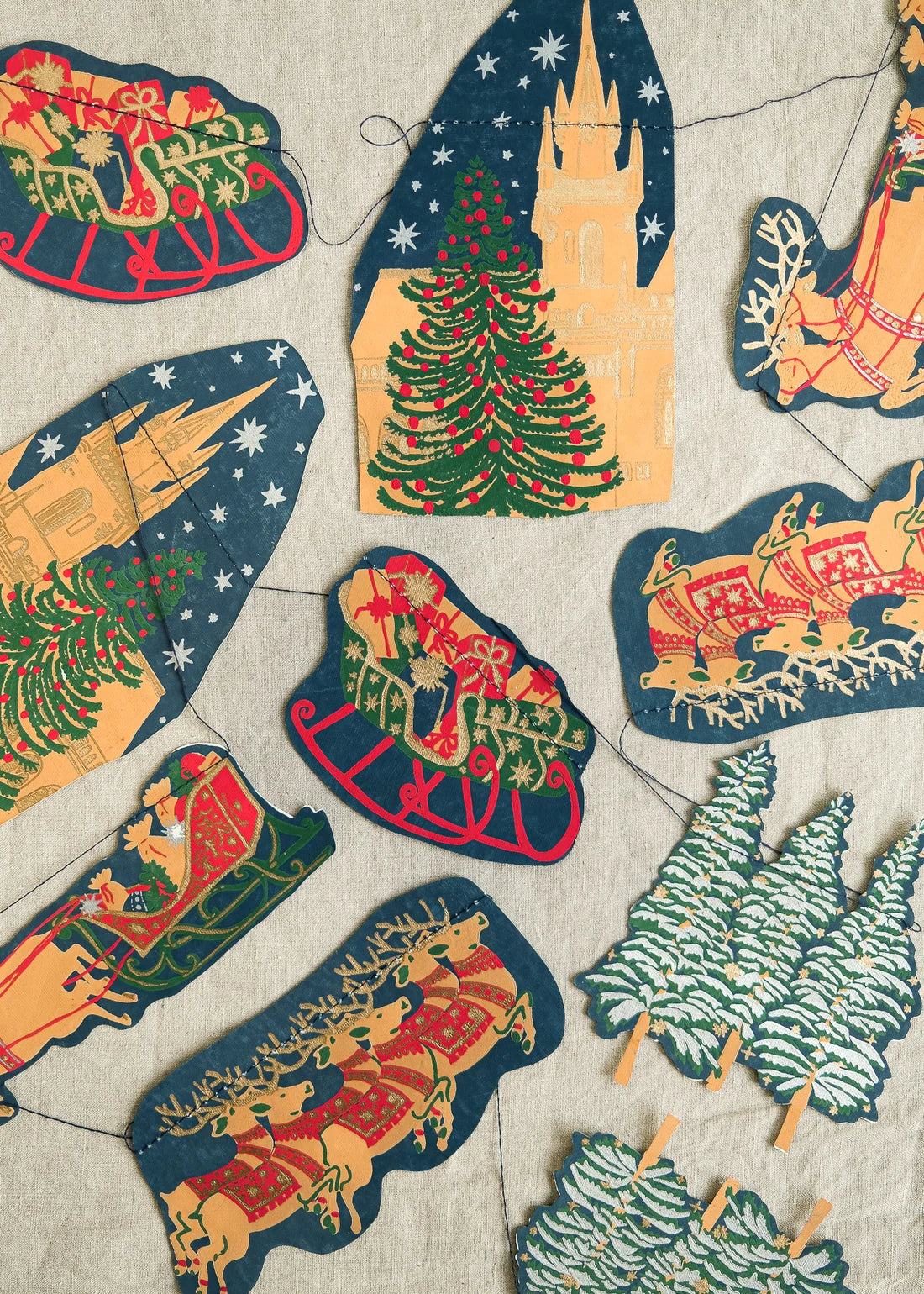 A festive Christmas garland made of multiple  paper cutouts on a string, featuring Santa in sleighs, Christmas trees, a castle, and holiday scenes, arranged on a light beige textured fabric background.