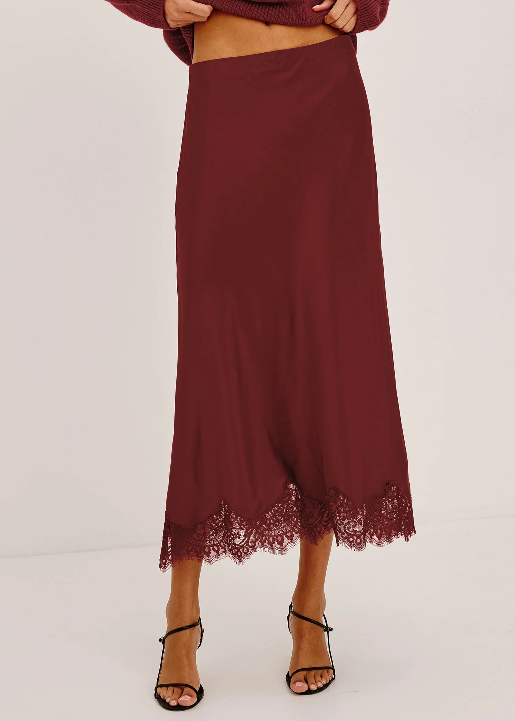 A model wearing a long, dark burgundy midi skirt with a lace hem, paired with black strappy heels and a matching burgundy sweater against a plain white studio background.