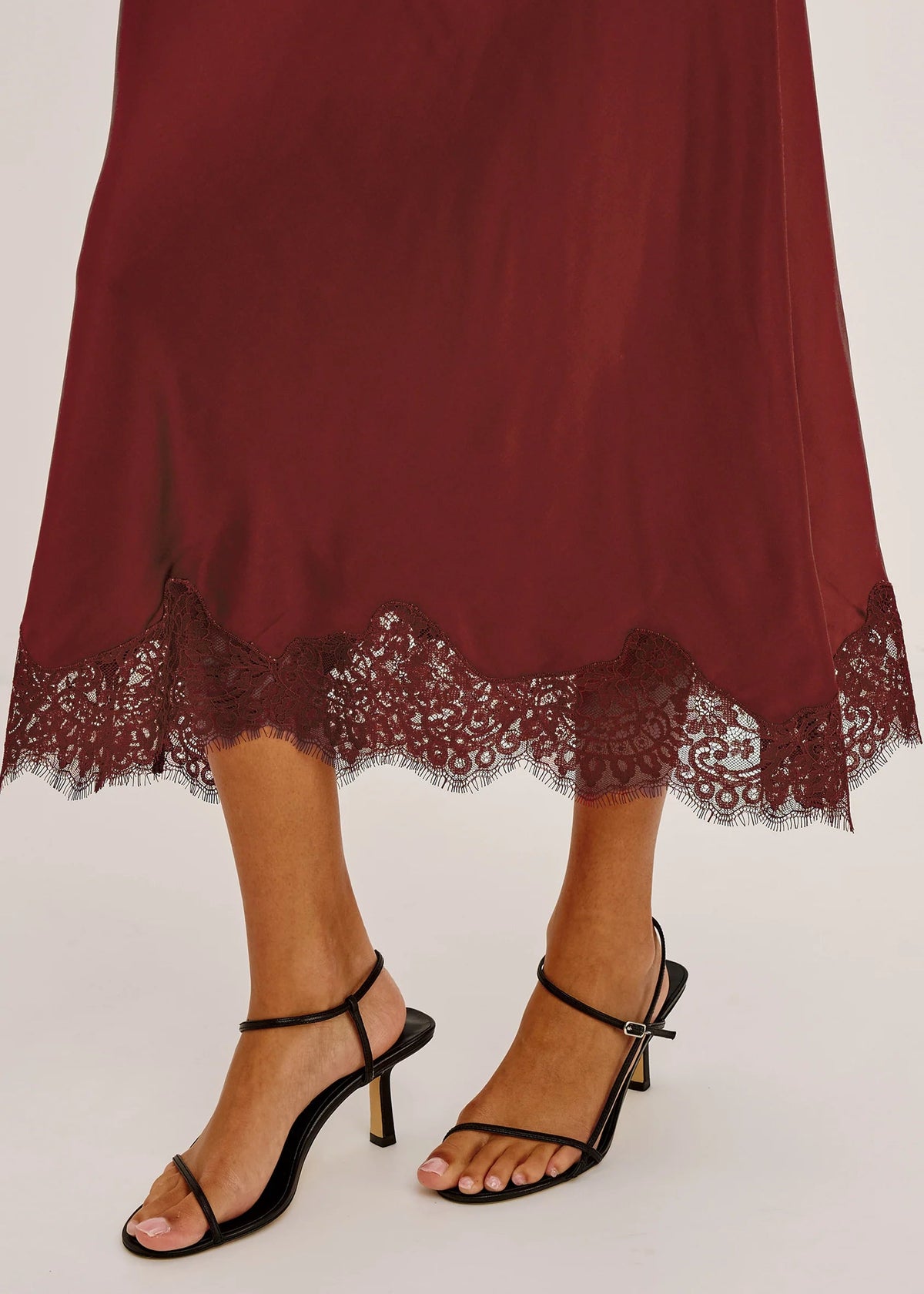 A close-up of a person's feet wearing black strappy high-heeled sandals with a burgundy midi skirt featuring a lace hem, set against a plain light gray background.