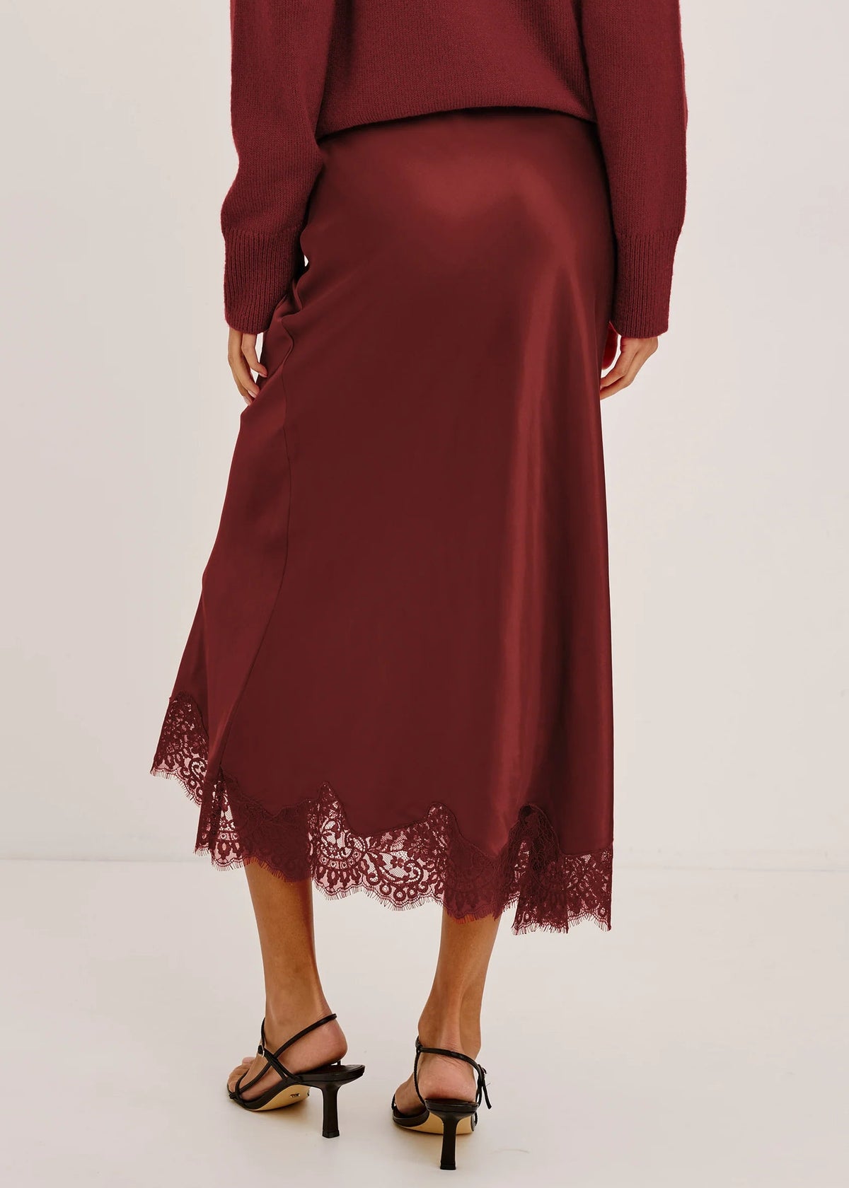 A woman wearing a long-sleeve satin midi skirt in deep burgundy with a lace hem, paired with black strappy heels, shown from the back against a plain light background.