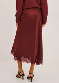 A woman wearing a long-sleeve satin midi skirt in deep burgundy with a lace hem, paired with black strappy heels, shown from the back against a plain light background.