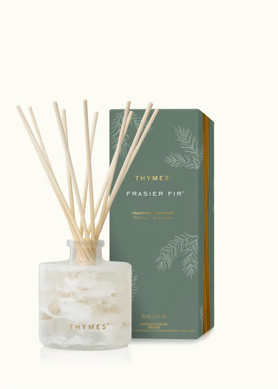 Thymes Frasier Fir reed diffuser with a clear glass bottle, white wax, and natural wood reeds, shown next to its sage green and gold packaging box on a light cream background.