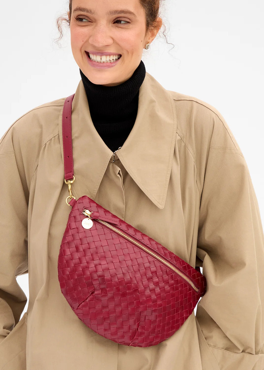 A woman wearing a beige trench coat over a black turtleneck, carrying a burgundy woven leather crescent-shaped crossbody bag with gold hardware.