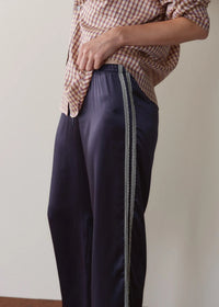 A person wearing dark navy wide-leg satin pants with a decorative braided stripe down the side, paired with a brown and white checkered shirt, shown from the waist down against a plain beige wall.